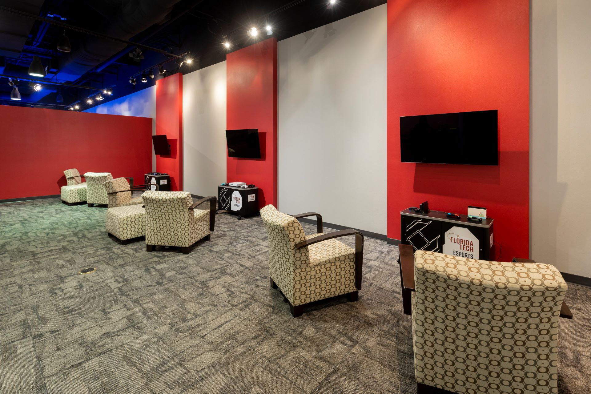 Interior of an Esports center with several seating areas featuring comfortable armchairs, each facing a wall-mounted television, against a backdrop of red and gray walls